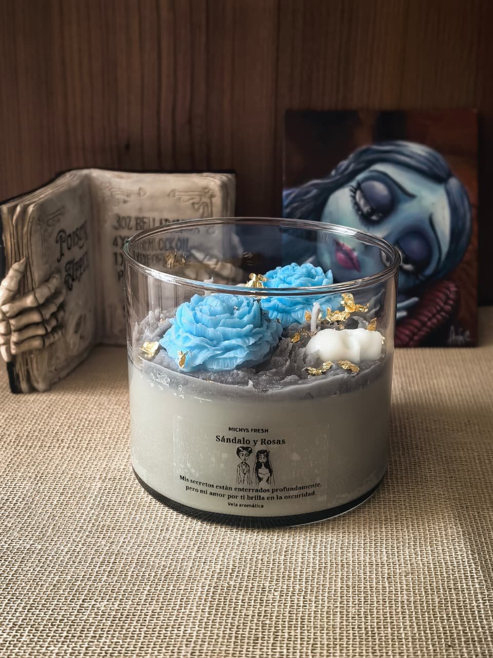 Vela aromática temática de Emily decorada con rosas azules y detalles dorados, acompañada de un libro de pociones y una ilustración al fondo.