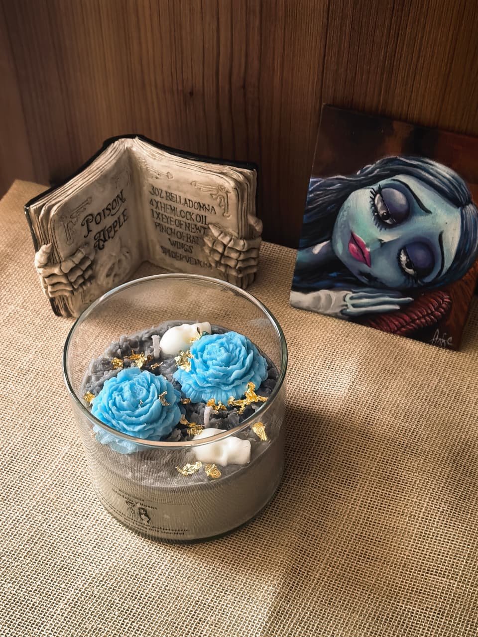 ela aromática de Emily en vaso de vidrio posicionada junto a un libro de hechizos abierto y un cuadro decorativo de la personaje.